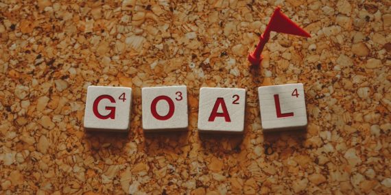 Scrabble tiles forming the word 'goal' with a small red flag on a cork board background.